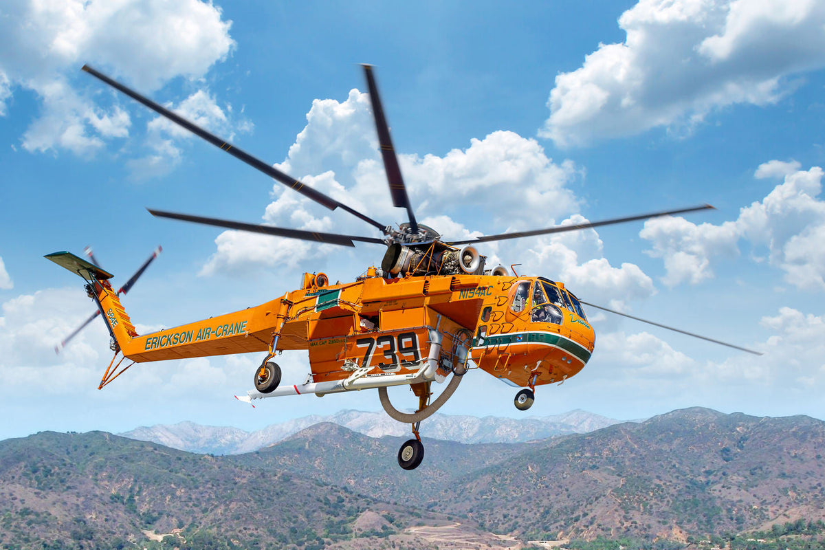 An orange and black rescue helicopter flying over a mountainous landscape under a blue sky with white clouds. The helicopter has 'EMERGENCY AIR SERVICE' printed on the side and the number '739' on the fuselage.