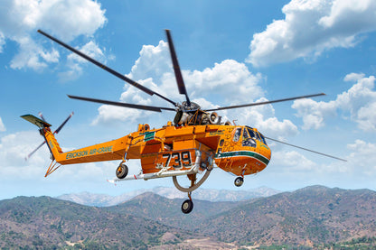An orange and black rescue helicopter flying over a mountainous landscape under a blue sky with white clouds. The helicopter has 'EMERGENCY AIR SERVICE' printed on the side and the number '739' on the fuselage.