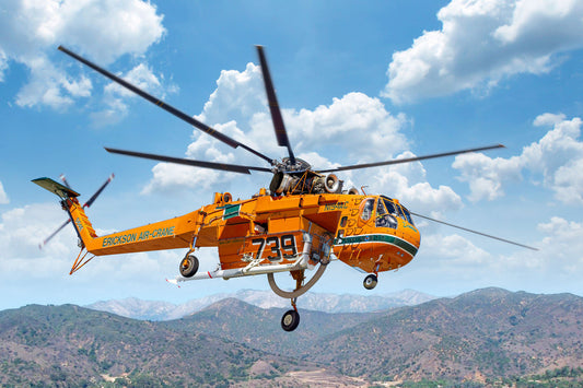 An orange and black rescue helicopter flying over a mountainous landscape under a blue sky with white clouds. The helicopter has 'EMERGENCY AIR SERVICE' printed on the side and the number '739' on the fuselage.