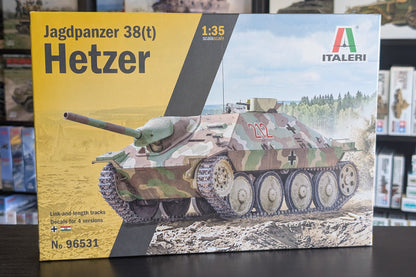 Front of an Italeri Jagdpanzer 38(t) Hetzer model kit box, shown standing on a dark tabletop with shelves of other boxes in the background. The packaging features a large yellow panel on the left, a dark diagonal band, and a printed illustration of the tank in camouflage on the right.