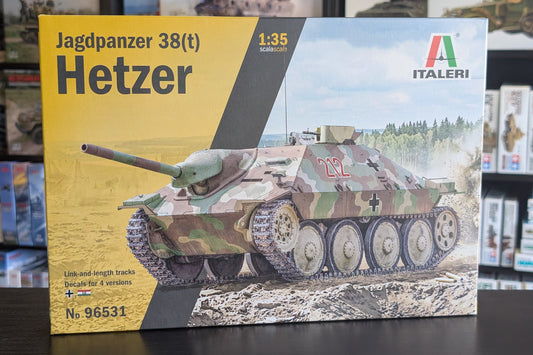 Front of an Italeri Jagdpanzer 38(t) Hetzer model kit box, shown standing on a dark tabletop with shelves of other boxes in the background. The packaging features a large yellow panel on the left, a dark diagonal band, and a printed illustration of the tank in camouflage on the right.