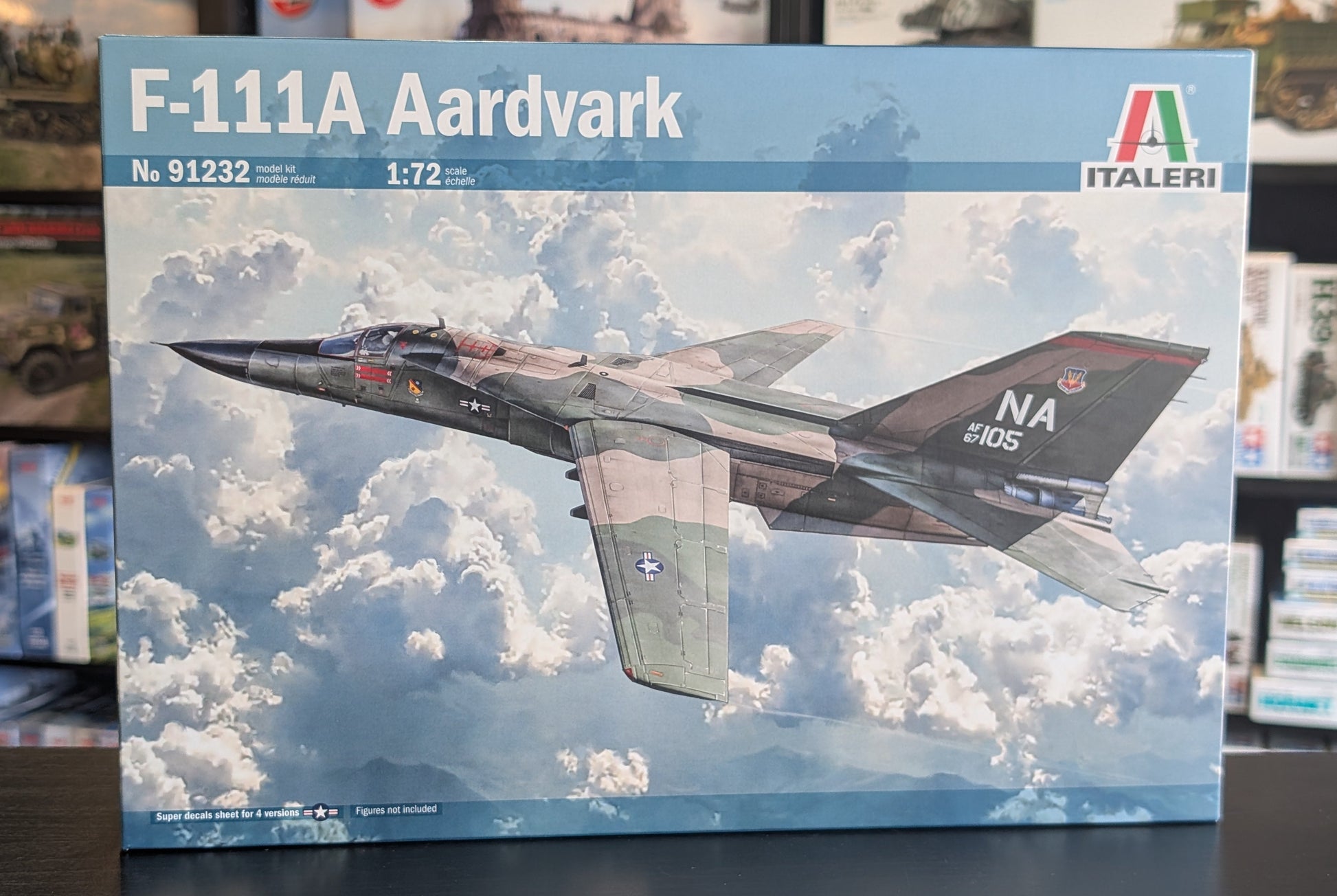 A boxed model kit for an F-111A Aardvark military fighter jet, shown upright on a dark tabletop with a blurred store or shelf background. The front of the box features a large printed photo of the jet flying through clouds, with the Italeri brand logo in the top right.