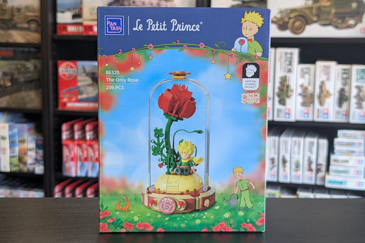 A boxed building block set for children titled 'Le Petit Prince' featuring a red rose under a glass dome, displayed on a dark tabletop with toy Pantasy - Le Petit Prince™ The Only Rose - 211 Pcs - Pa86320 blurred in the background.