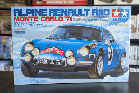 Tamiya Alpine Renault A110 Monte-Carlo '71 model car box front, showing a blue rally car with sponsor decals on a snowy road, photographed in front of a blurred shelf background.