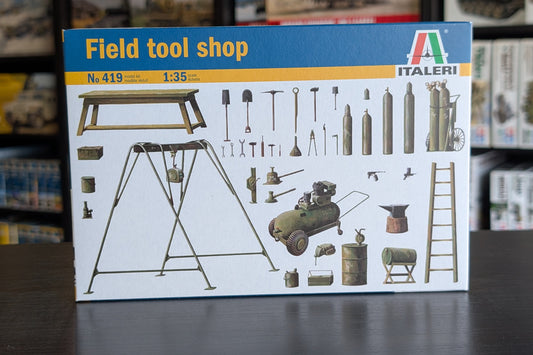 A boxed model kit labeled 'Field tool shop' by Italeri, shown standing on a dark tabletop with blurred shelves of other boxes in the background. The front of the box features a blue header, yellow banding, and illustrated tools and equipment for a model workshop.
